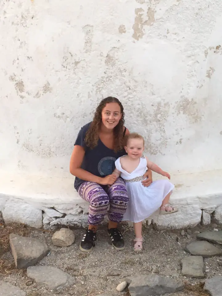 The founder of island hopping in Greece sitting at the base of a Mykonos windmill with her daughter wearing traditional Greek clothing.