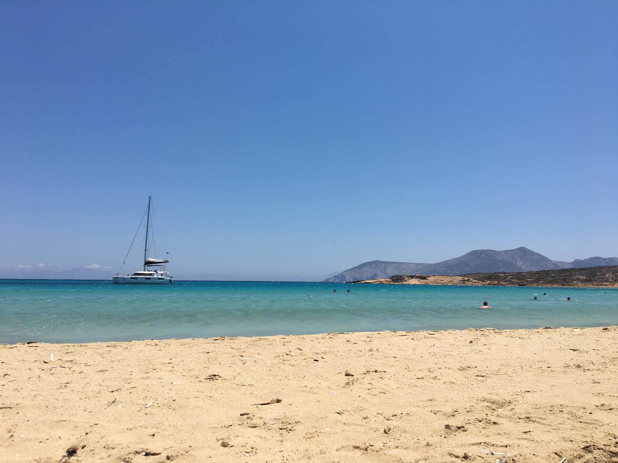 A serene beach scene on Koufinisi island featuring a catamaran yacht anchored in a clear blue bay with a sandy beach in the foreground. The background displays hilly landscapes under a bright, cloudless sky, and several people are swimming in the calm sea.







