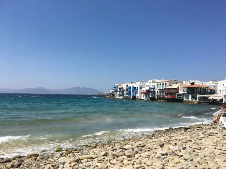 Mykonos Town viewed from the rocky shore, showcasing a vibrant seascape with traditional white buildings and blue accents, famous for its picturesque charm.