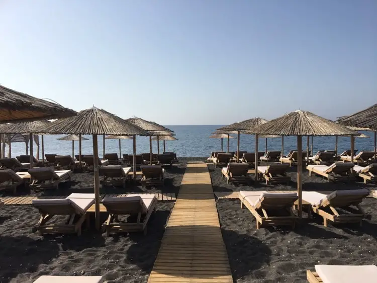 Kamari beach in Mykonos with black sand. Lines of sunbeds leading to the water
