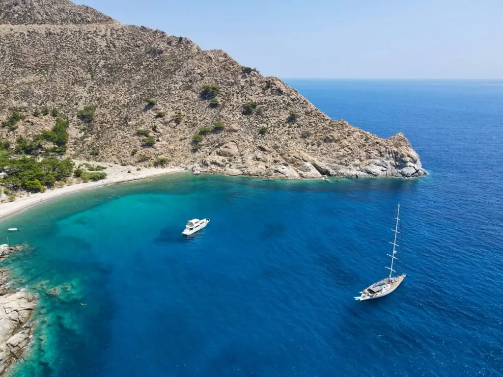 Secluded cove on a Greek island showcasing a tranquil beach with crystal-clear turquoise waters, surrounded by rugged cliffs and lush greenery, ideal for private yacht anchoring and relaxation.