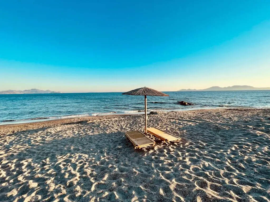 greek island hopping route, Secluded sandy beach with a single straw umbrella and wooden lounger, facing a serene sea under a clear sky, offering a perfect spot for peaceful relaxation. The gentle waves and distant mountain silhouette enhance the tranquil atmosphere.