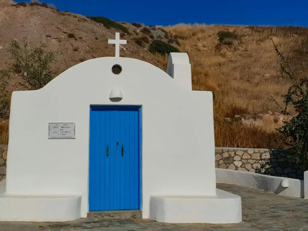 Charming white chapel with bright blue door set against a rocky hillside in Anafi, Greece.